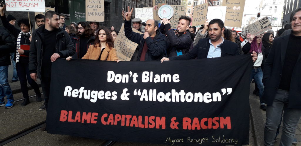 Een groep demonstranten. Voorop dragen enkele mensen een spandoek met de tekst: Don't blame refugees & "Allochtonen". Blame capitalism and racism. Migrant refugee solidarity.