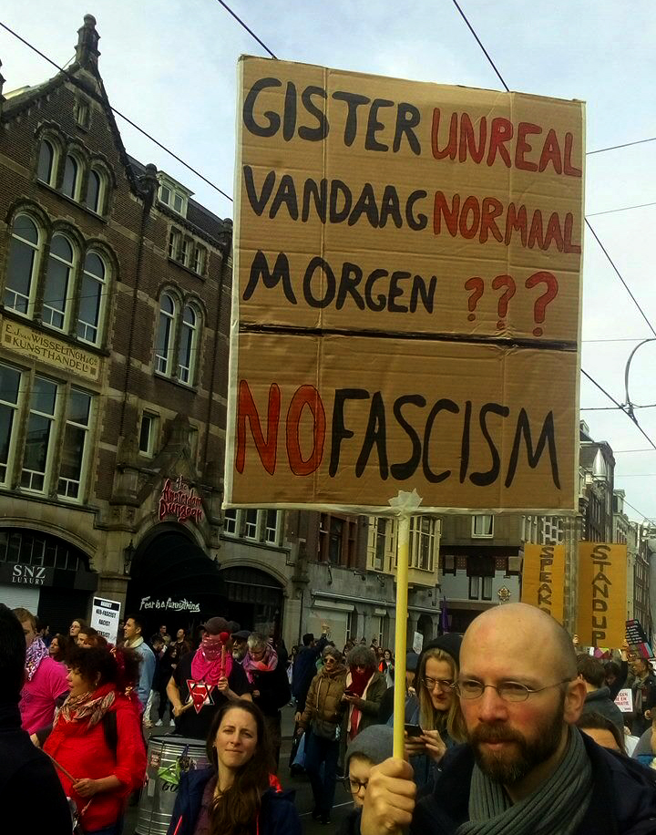 Een persoon met een bril en een baard houdt een kartonnen bord vast met daarop de tekst "Gisteren unreal - vandaag normaal - morgen ??? / No fascism".