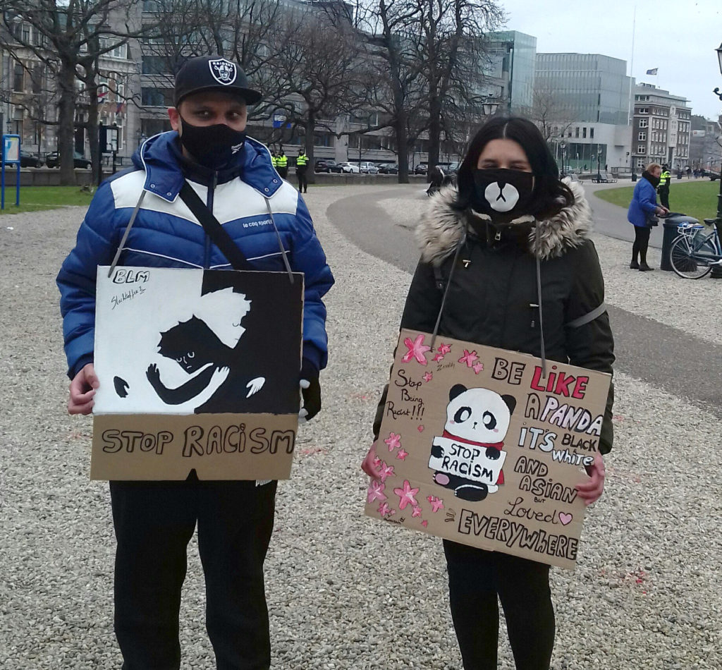 Twee jonge mensen bij de demonstratie in Den Haag. Kantoorgebouwen op de achtergrond. Politiemensen in de verte. De ene persoon draagt een bord waar "BLM - stop racism" op staat. De andere persoon draagt een bord waar op staat "Stop being racist!!! Be like a panda - it's black, white and Asian, but loved everywhere". Op het bord is een panda getekend die een bordje vasthoudt met daarop "stop racism".