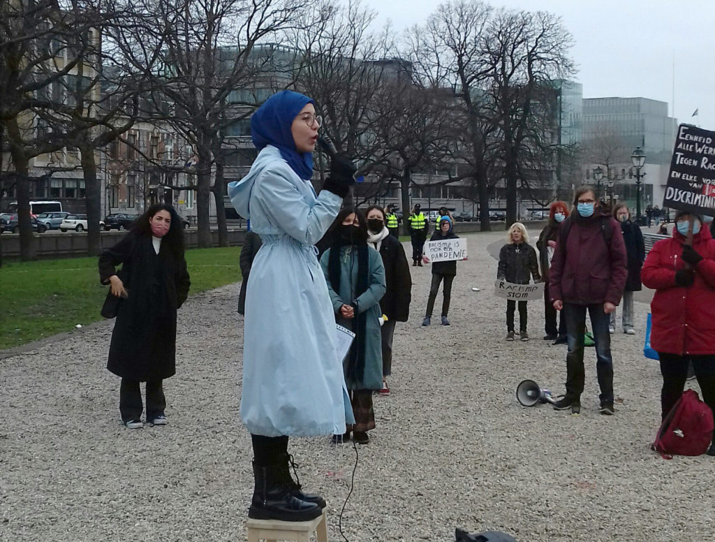 Een persoon met een lichtblauw lange jas en een blauwe hijab spreekt via een microfoon. Om de persoon heen staan een aantal toehoorders, een deel met protestborden. Op de grond ligt een megafoon.