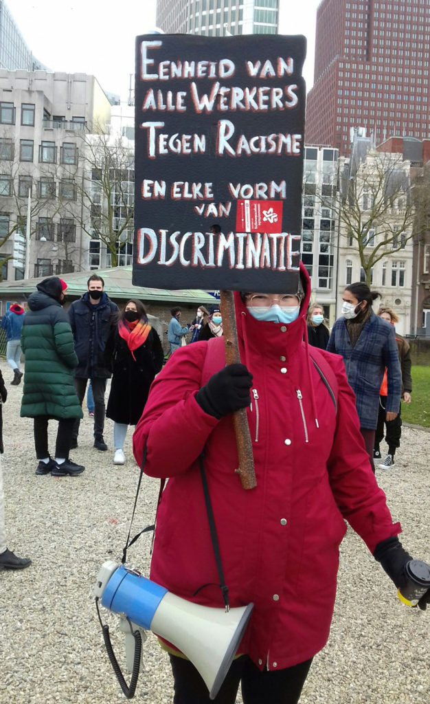 Een warm ingepakte persoon met een rode jas aan en de capuchon over het hoofd met een bord met de tekst: "Eenheid van alle werkers tegen racisme en elke vorm van discriminatie".