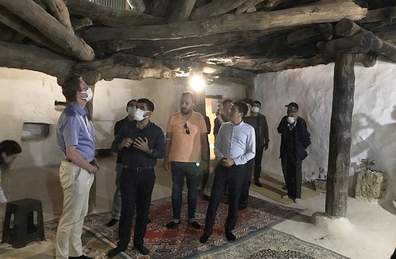 Een groep mannen met mondkapjes staat in het oude cemhuis. Het plafond is van oude houten balken. De muur is onregelmatig van structuur en witgekalkt. Op de lemen vloer liggen tapijten. Tegen de wand twee stenen wasbakjes.