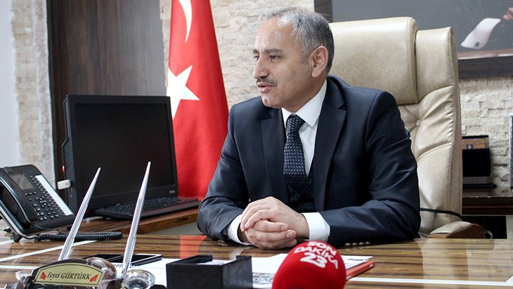 Feyzi Gürtürk achter zijn glimmende houten bureau, op een grote leren bureaustoel. Hij draagt een donkerblauw pak en dito stropdas. Op de achtergrond de Turkse vlag.