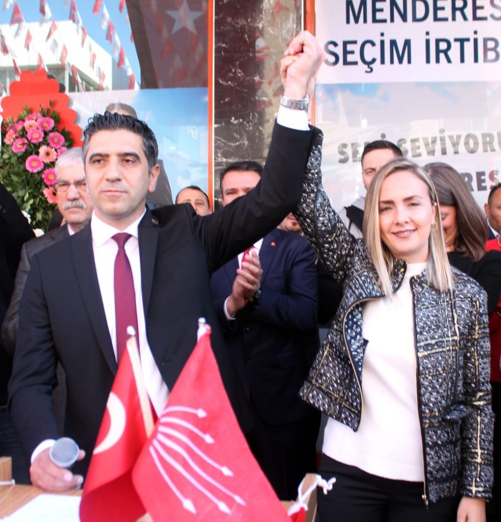 Mustafa Kayala, donker pak met rode stropdas, houdt de hand omhoog van een persoon met lange blonde haren en een jasje met gouden strepen erin. Op de achtergrond klappende mensen.