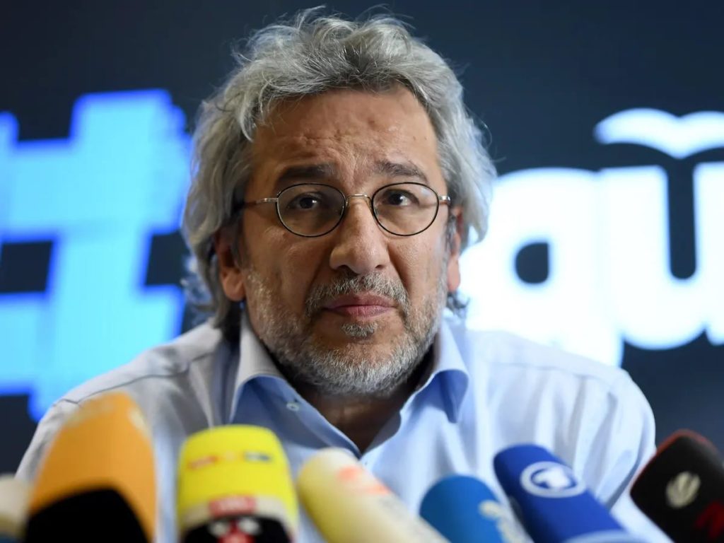 Foto van Can Dündar met meerdere nieuwsmicrofoons voor zich.