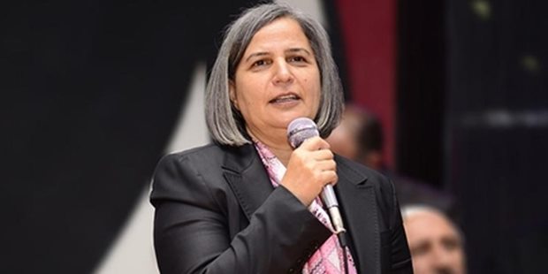 Gültan Kışanak die in een microfoon spreekt. Ze heeft grijze haren in een lange boblijn. Ze draagt een zwart jasje en een roze geruite sjaal.