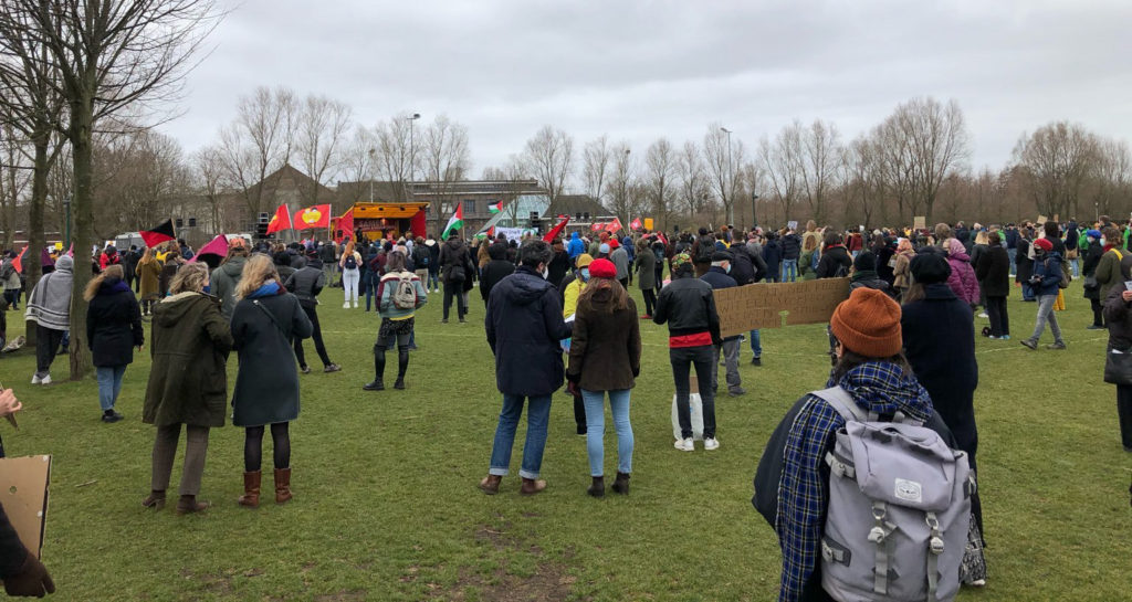 Een grasveld met een menigte demonstranten erop. Vlaggen van Palestina, anarchismevlaggen.