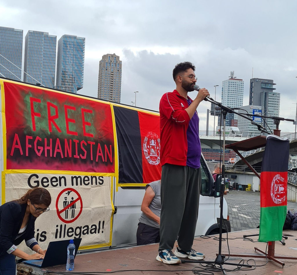 Akef aan het woord op het podium. Akef draagt een grijze joggingbroek, een paars shirtje en een rood vest. Hij heeft zwarte krullen en een baardje. Op de achtergrond de hoogbouw van Rotterdam.