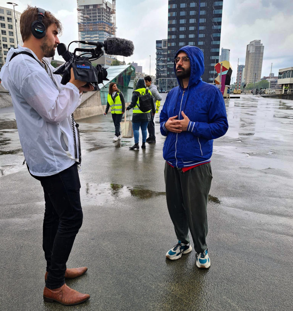 Akef wordt geïnterviewd. Het regent en Akef heeft zijn capuchon opgedaan. De journalist draagt een witte regenjas en filmt Akef.