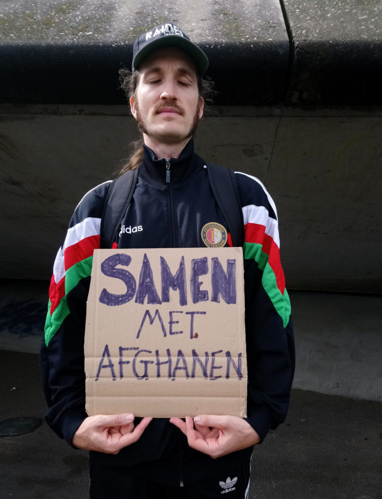Een demonstrant in een Feyenoord-jasje draagt een kartonnen protestbord met de tekst 'Samen met Afghanen'.
