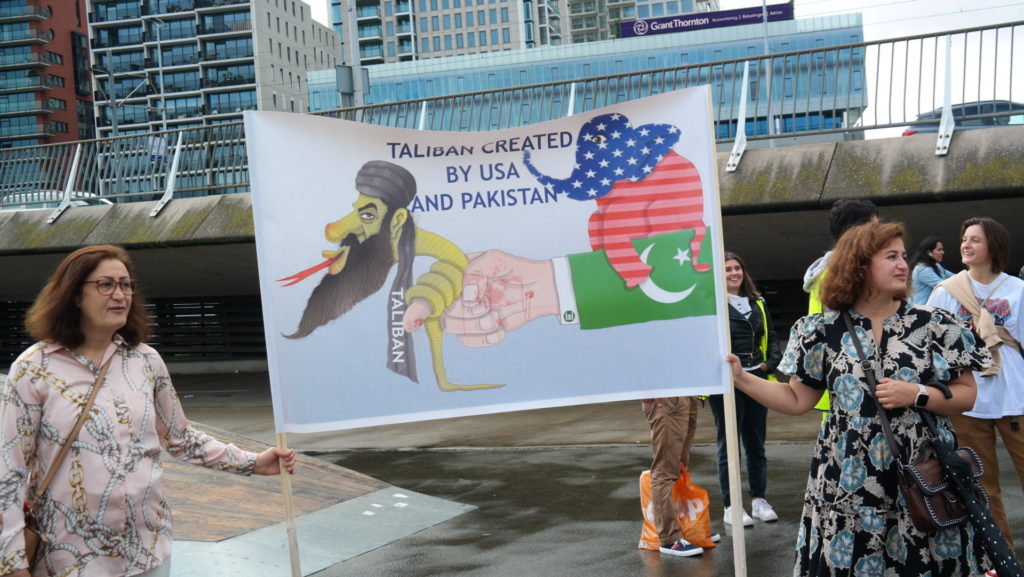 Een spandoek met daarop een spotprent van een Taliban-strijder in de vorm van een slang, die om een bloedende vinger gewikkeld zit. De hand zit aan een arm met de Pakistaanse vlag. Op de arm zit een Amerikaanse olifant.