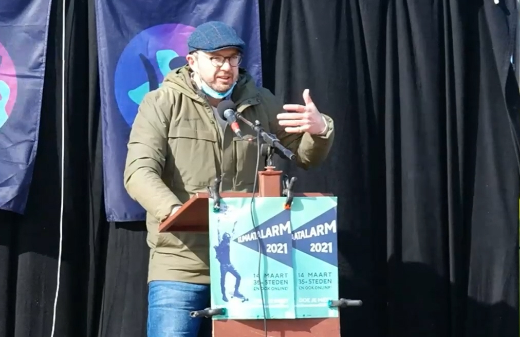 Foto van Christian Henderson die achter een lessenaar een speech geeft bij het Klimaatalarm in Leiden. Christian draagt een tweed pet en een legergroene warme jas. Op de lessenaar hangt de mintgroene poster van het Klimaatalarm 2021.