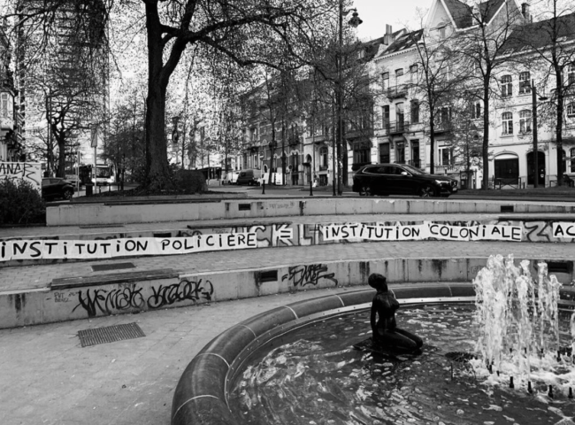 Een fontein op de voorgrond met een vrouwelijk naakt erin. Op de achtergrond een trap waarop de tekst "Institution policiere / institution coloniale" staat.