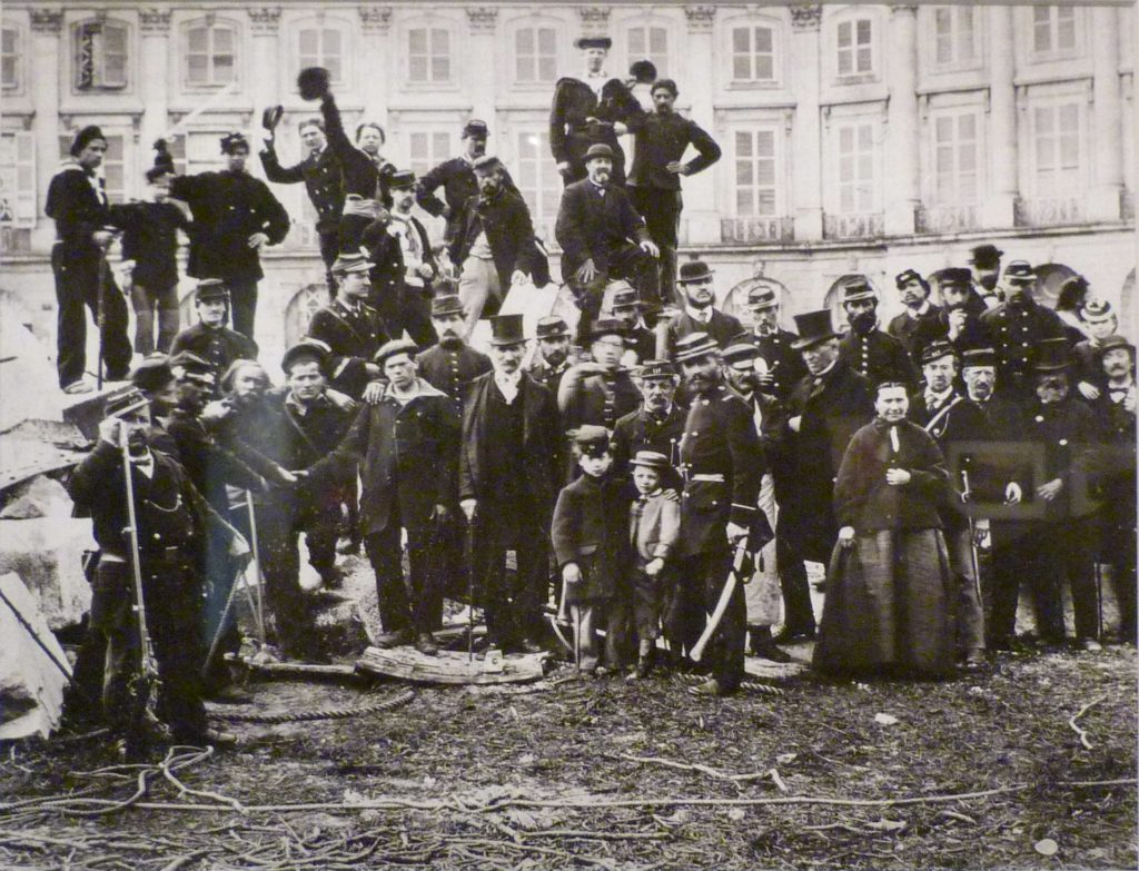 Een poserende groep deelnemers aan de Commune van 1871. Iedereen draagt donker gekleurde kleding. Op de voorgrond twee kinderen en een vrouw met een wijde rok. Achteraan staan mensen op een verhoging. Sommigen nemen hun pet af voor de foto.