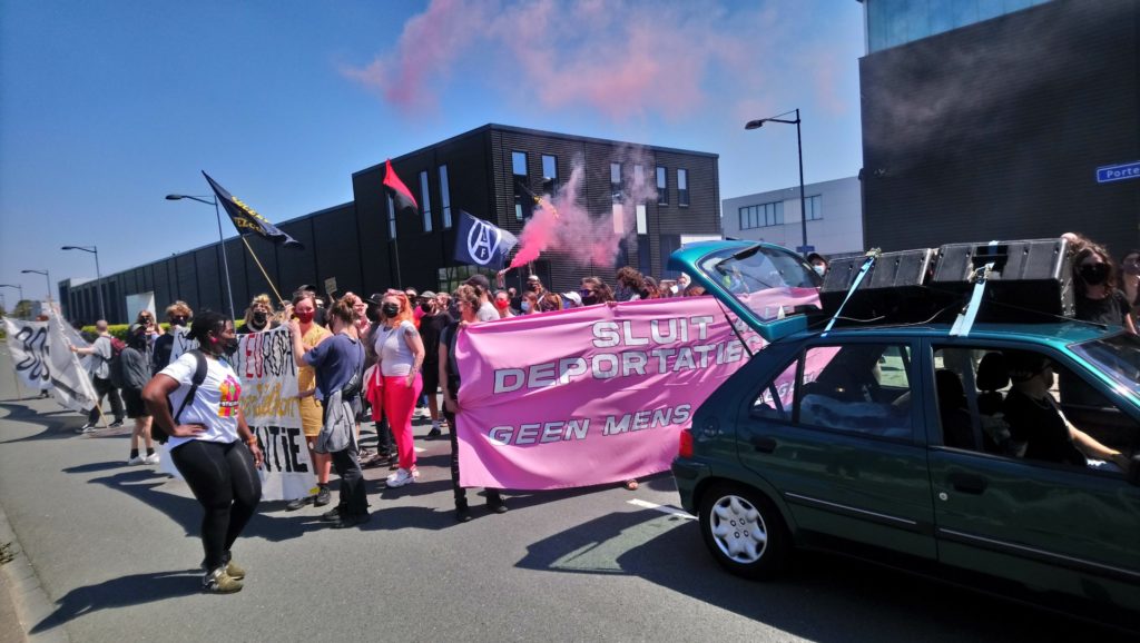 Overzicht van de demo, met vooraan een auto met geluidsinstallatie. Een roze flare is net afgestoken. Iemand draagt een roodzwarte anarchistische vlag aan een lange stok.