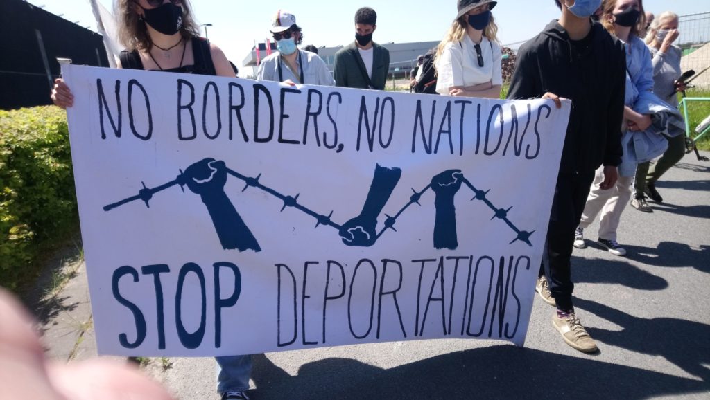 Spandoek met de tekst "No borders - no nations - stop deportations". Erop afgebeeld drie vuisten die prikkeldraad kapot trekken.