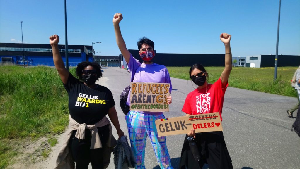 Drie demonstranten met de vuisten in de lucht. Eentje draagt een "Gelijkwaardig BIJ1" shirt, eentje heeft een bord met de tekst "Refugees are my G's - open the borders" en eentje draagt een shirt met de tekst "Stop war not people" en een bord met de tekst "Gelukzoekers" waarbij 'zoekers' is doorgestreept en vervangen door 'delers' met een hartje erachter.