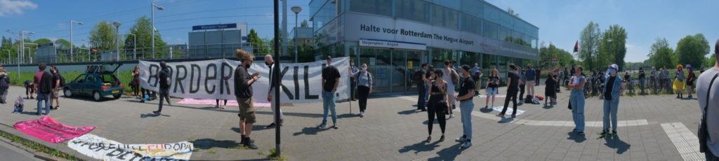 Breedbeeldfoto met daarop het enorme spandoek "BORDERS KILL" en een deel van de demonstranten.