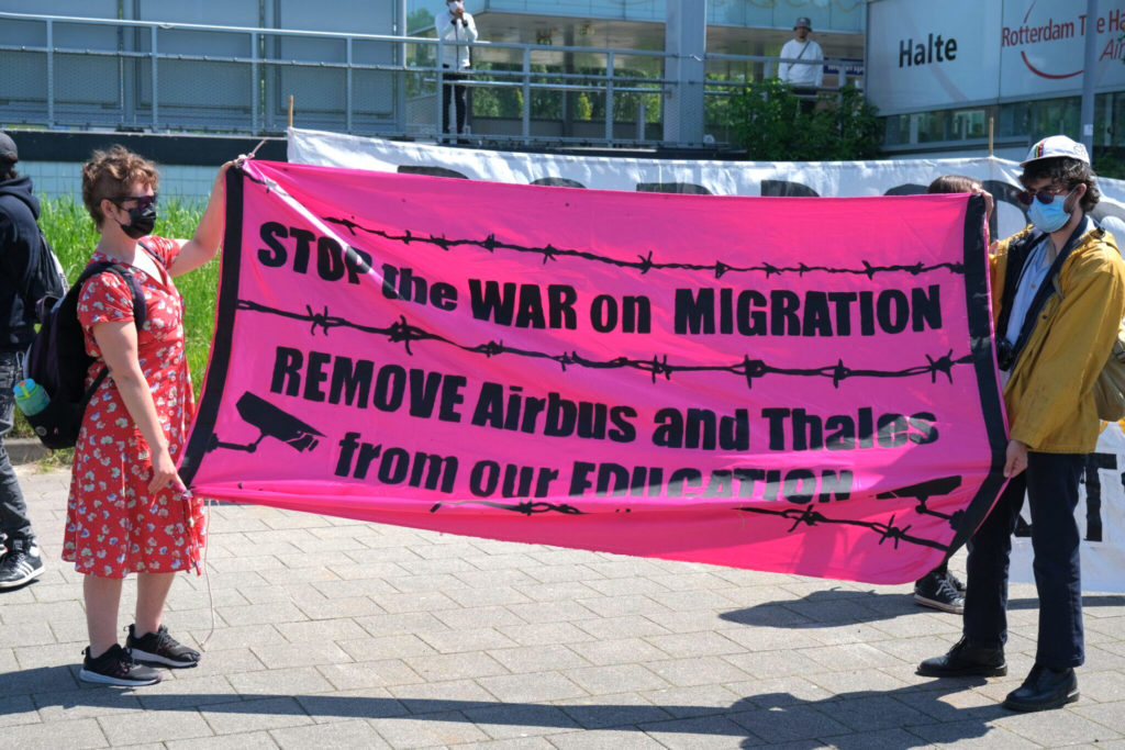 Twee deelnemers met een knalroze spandoek met de tekst "Stop the war on migration - remove Airbus and Thales from our education".