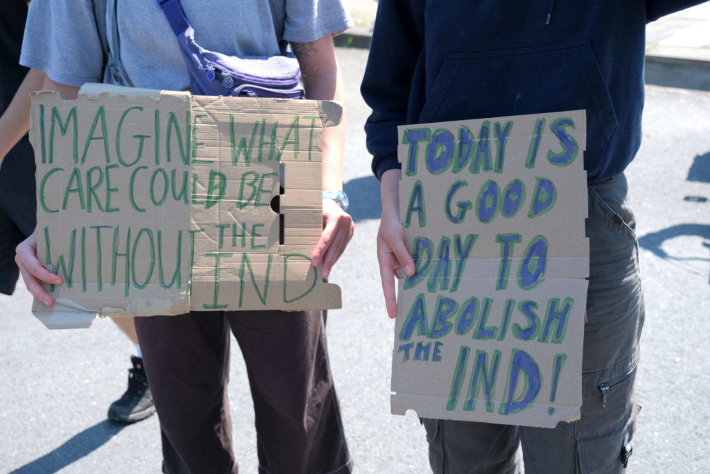Twee borden met de teksten "Imagine what care could be without the IND" en "Today is a good day to abolish the IND!".