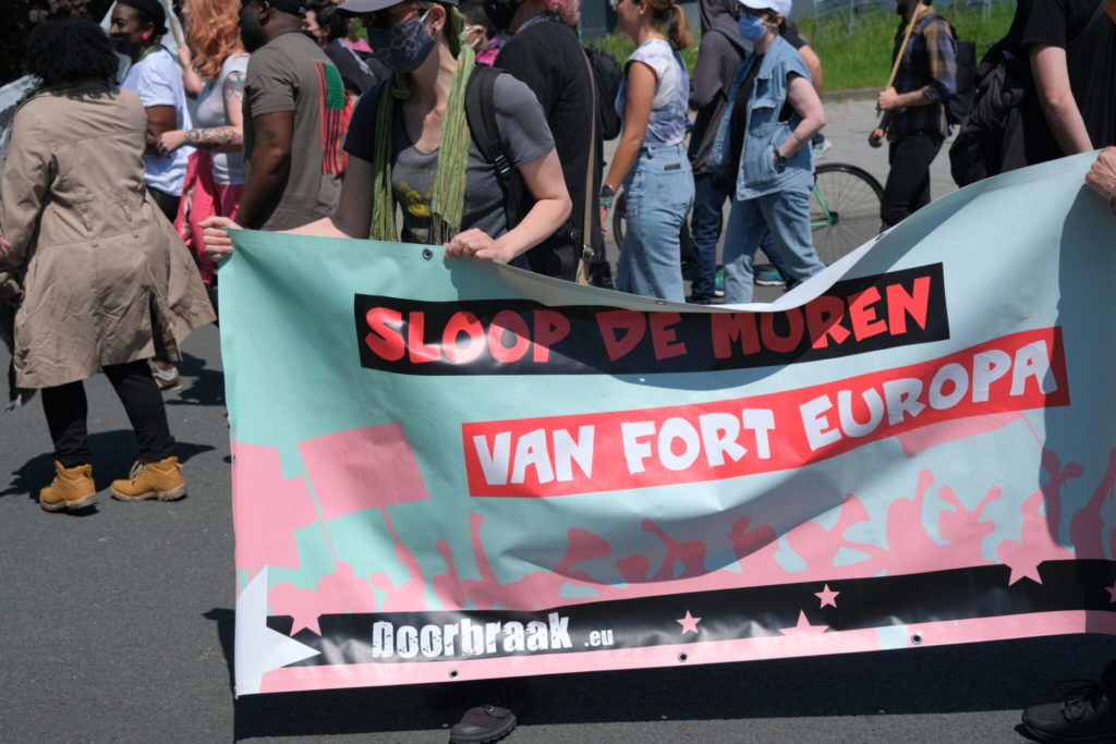 Doorbraak-spandoek in mintgroen en zachtroze met de tekst "Sloop de muren van Fort Europa".