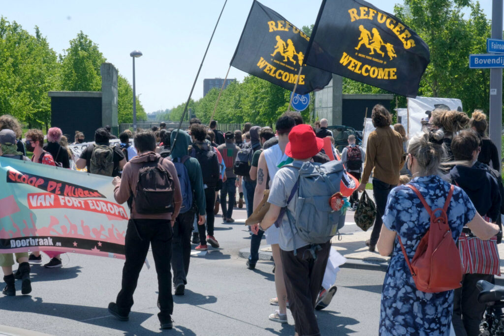 Overzicht van de deelnemers. Twee mensen dragen "Refugees welcome" vlaggen aan lange stokken.