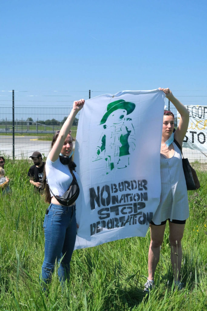 Twee mensen met een spandoek met daarop een beertje met een koffer en de tekst "No border no nation stop deportation".