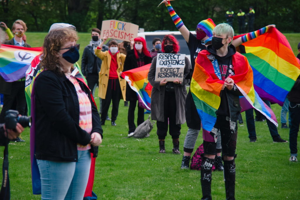 Op de achtergrond een activist met lange regenboog-armwarmers. Op de voorgrond iemand met de regenboogvlag om het lijf geslagen als een soort omslagdoek.