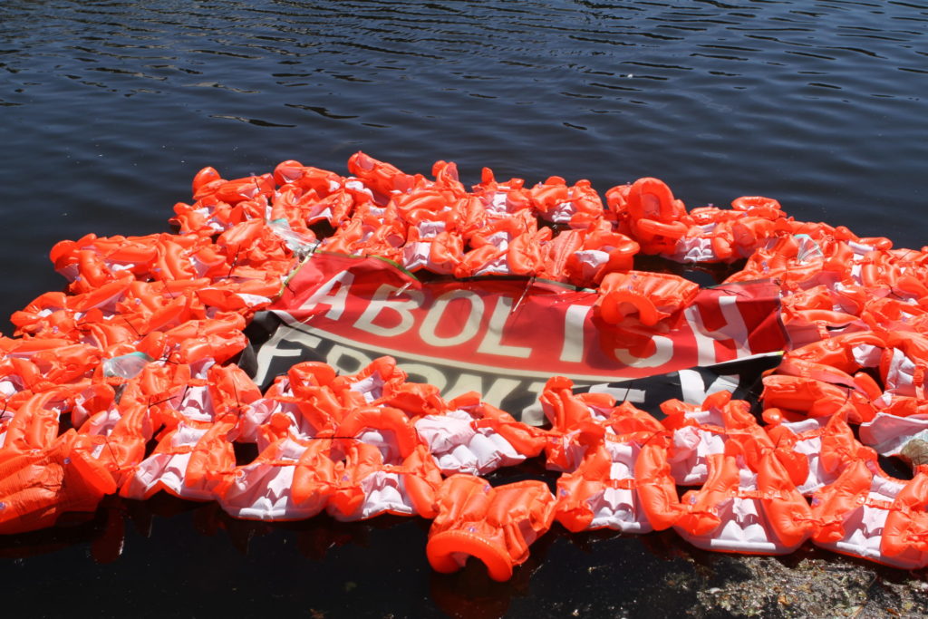 Een heleboel zwemvesten in de Hofvijver die een spandoek laten drijven met daarop de tekst "ABOLISH FRONTEX".