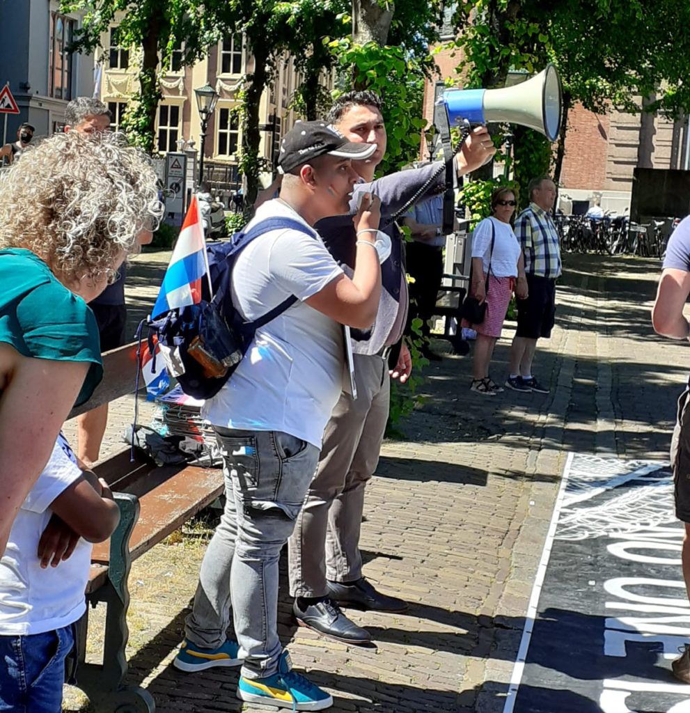 Een demonstrant met een rugzak op met een Nederlands vlaggetje eraan spreekt in de megafoon, die door iemand anders wordt vastgehouden.