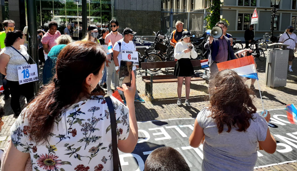 Een jonge demonstrant voert het woord en leest iets op van een smartphone. Omstanders dragen borden waarop te lezen is hoe lang ze al in Nederland zijn.