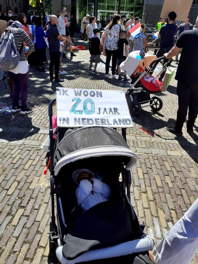 Een kinderwagen op de voorgrond, op de schaduwkap ligt een bord "Ik woon 20 jaar in Nederland". Een groep met demonstranten luistert naar een spreker die niet in beeld is.