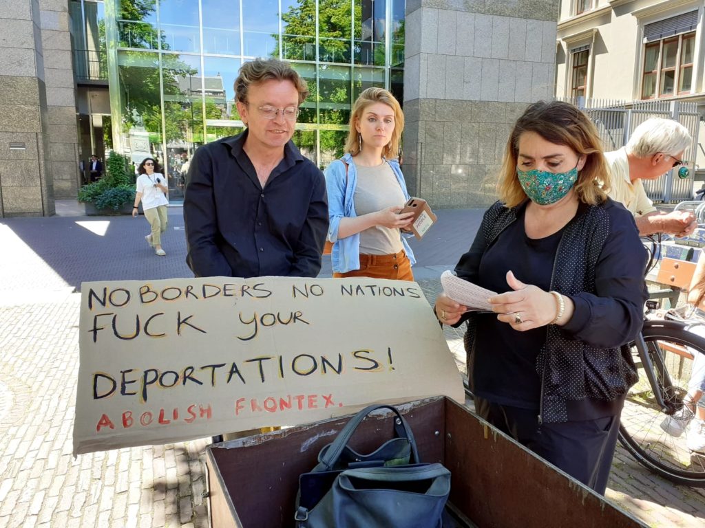 Fatima Faïd met een groen mondkapje met bloemen erop naast een bakfiets waarop een bord ligt met de tekst "No borders no nations, fuck your deportations! Abolish Frontex."