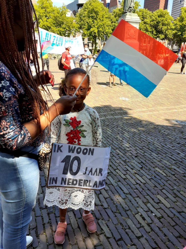 Een kind met een mooie bebloemde kanten jurk aan draagt een bord "Ik woon 10 jaar in Nederland". Naast het kind staat iemand met veel vlechtjes in het haar en een Nederlands vlaggetje in de hand.