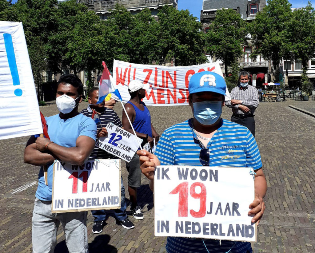 Twee demonstranten met hun mondkapjes op en borden die zeggen hoe lang ze in Nederland zijn (11 en 19 jaar).