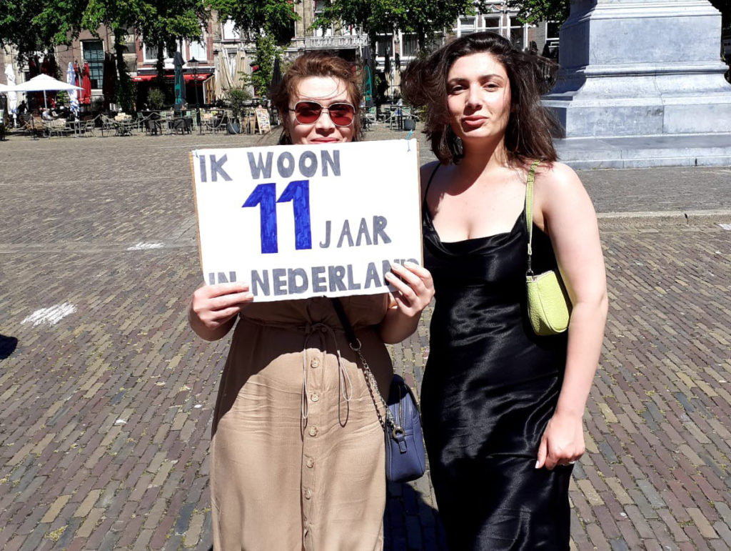 Twee mensen waarvan eentje in een zwarte satijnen jurk en de ander een bruine rok, deze draagt een bord "Ik woon 11 jaar in Nederland".