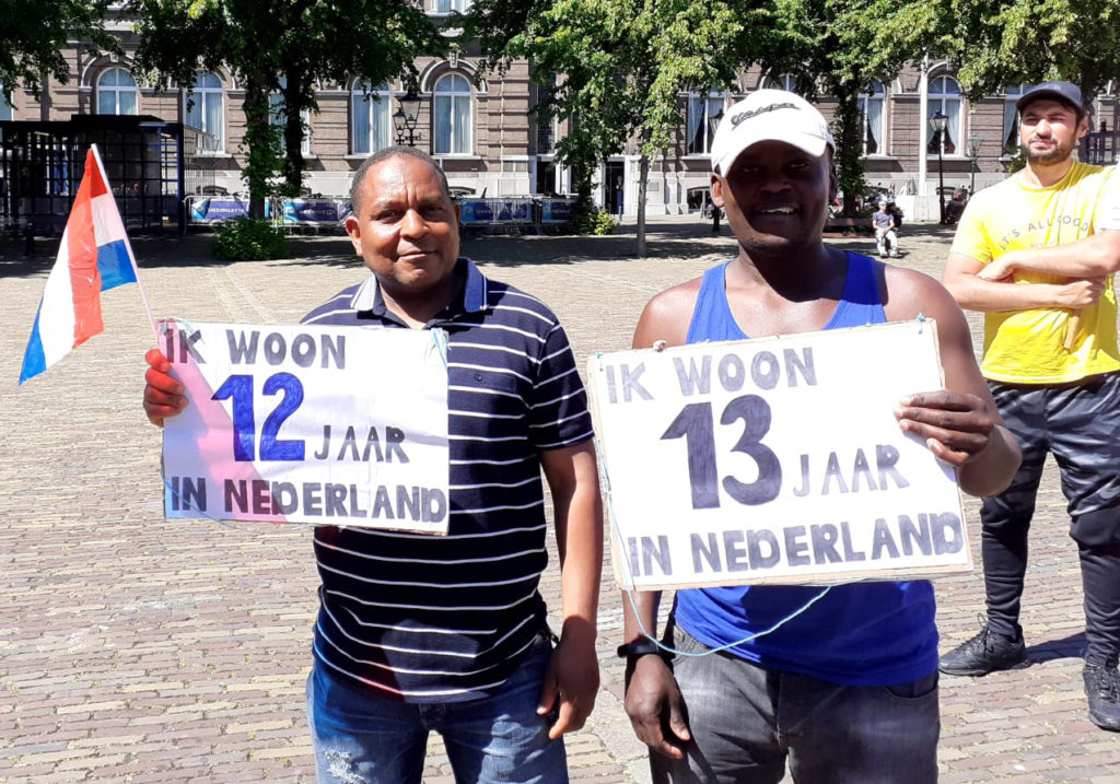 Twee mensen waarvan eentje in een blauw streepjesshirt en de andere in een blauw hemd met borden dat ze 12 en 13 jaar in Nederland zijn.