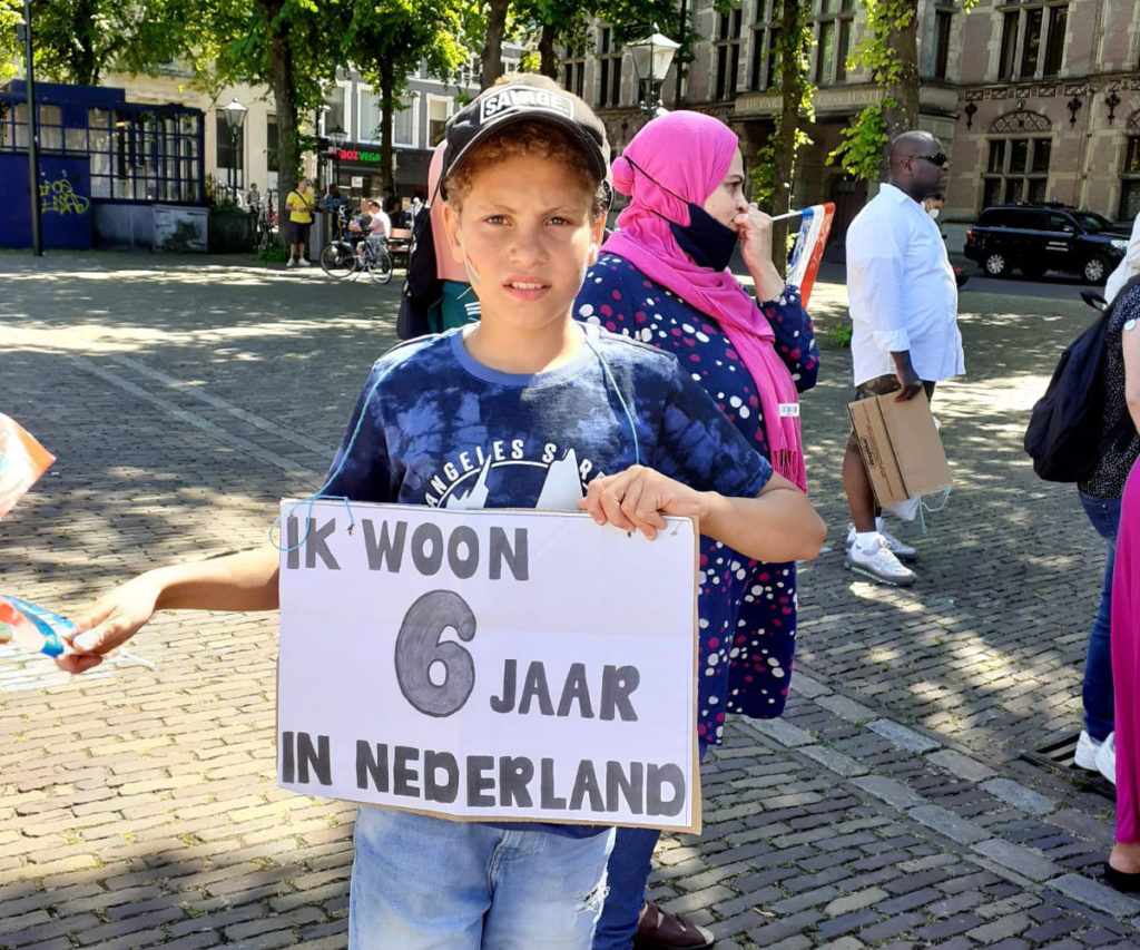 Een kind met een zwart petje op draagt een bord met de tekst "Ik woon 6 jaar in Nederland".