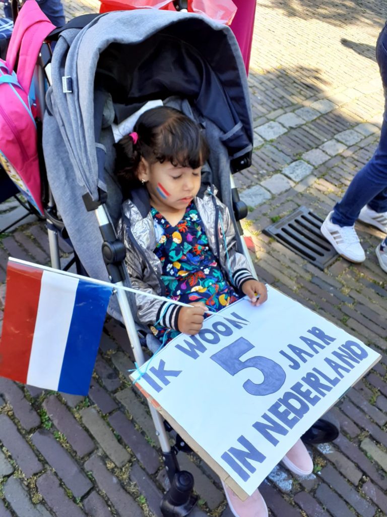 Een kindje is in slaap gevallen in de buggy , met een Nederlands vlaggetje in de hand en een bord op schoot met de tekst "Ik woon 5 jaar in Nederland".