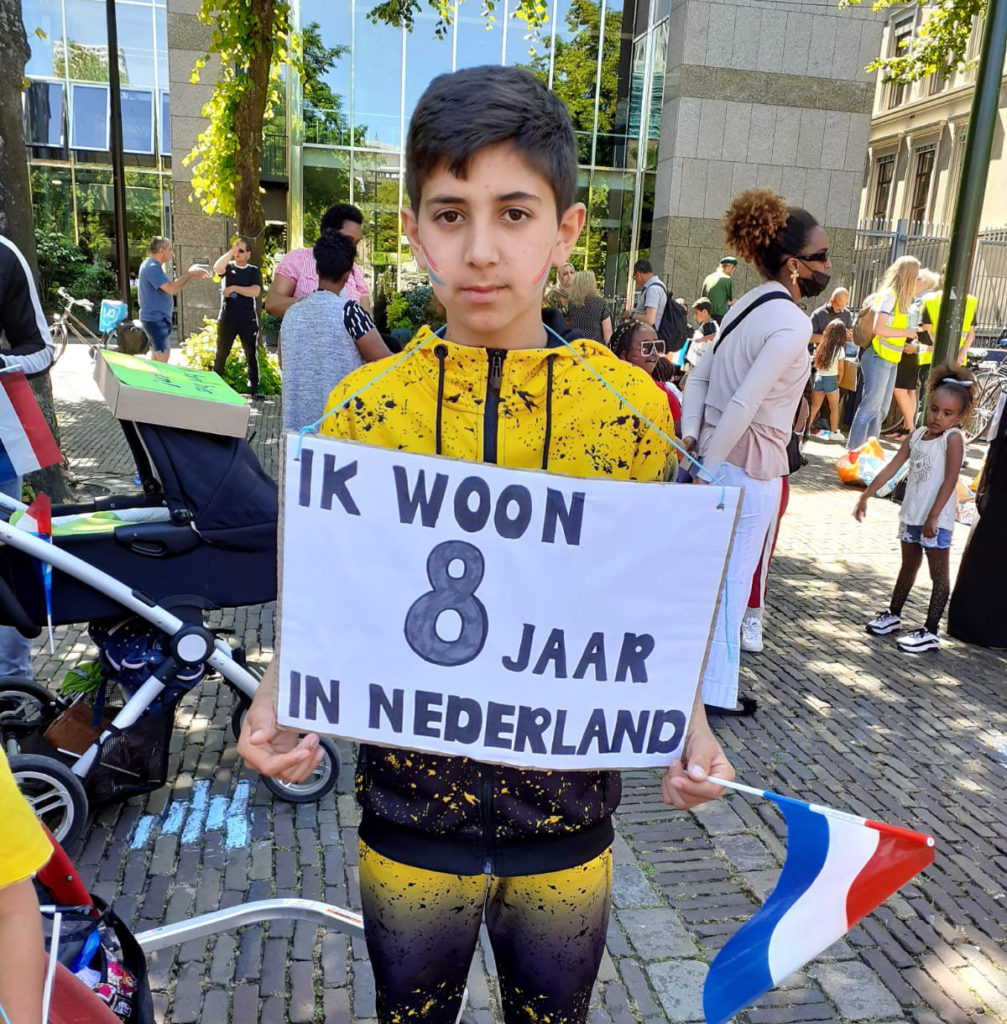 Een kind met een fel geelzwart trainingspak aan en Nederlandse vlaggetjes op de wangen. Op zijn bord staat "Ik woon 8 jaar in Nederland".