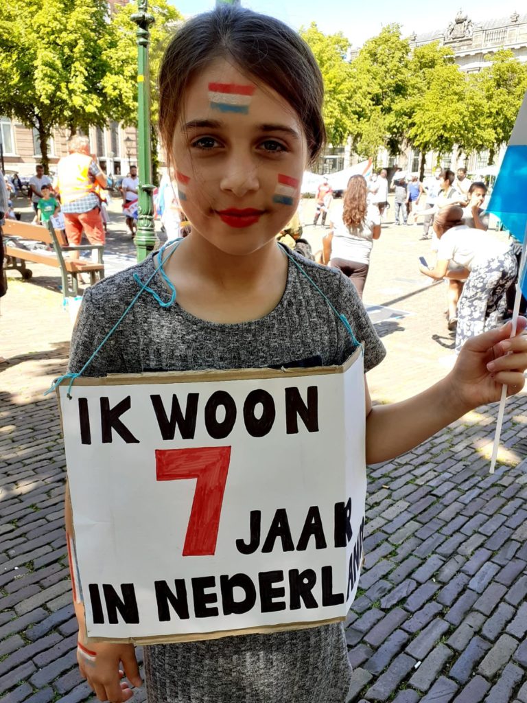 Een kind dat vriendelijk in de kamer kijkt. Ze heeft drie Nederlandse vlaggetjes op haar gezicht geschminkt staan. Ze draagt een bordje met "Ik woon 7 jaar in Nederland".