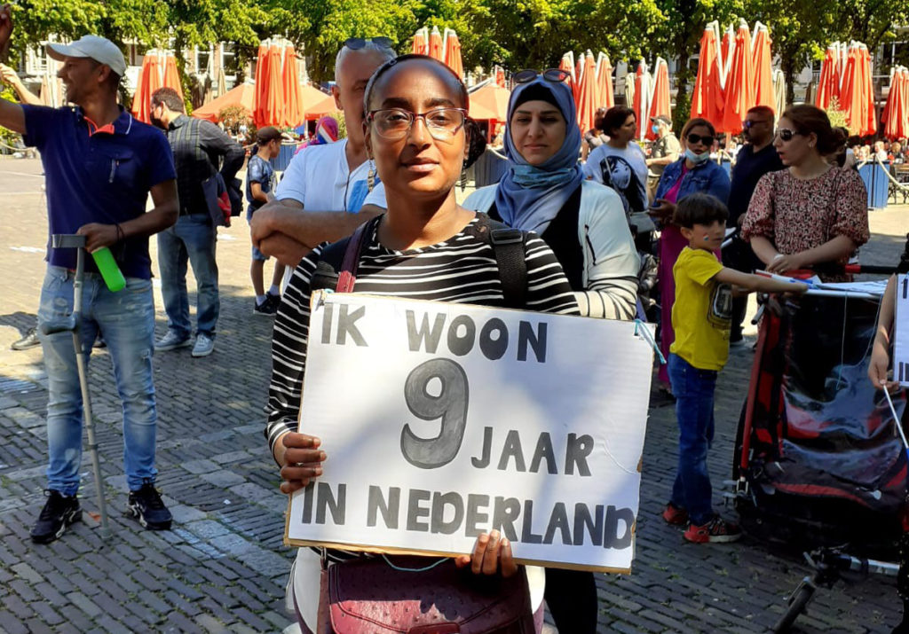 Iemand met een zwartwit streepjesshirt en een rode tas met een bord "Ik woon 9 jaar in Nederland".