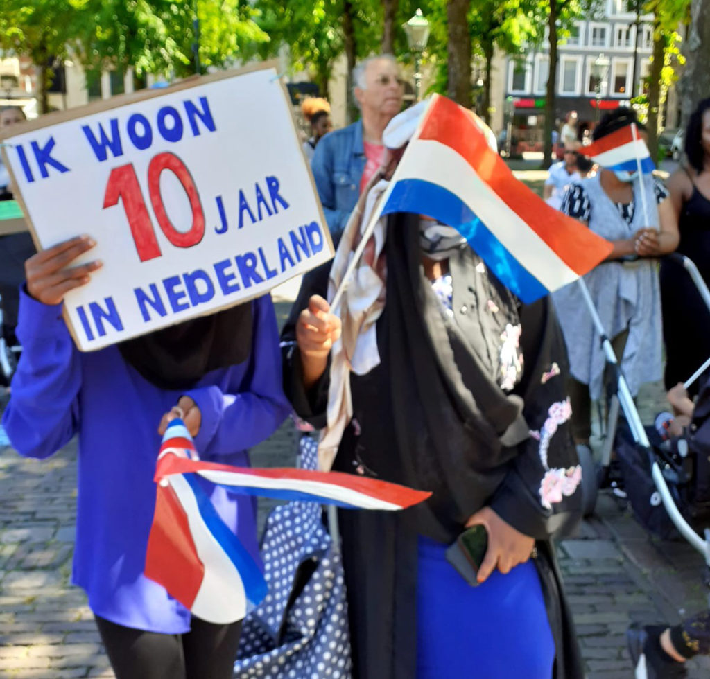 Twee mensen van wie de gezichten wegvallen achter hun vlaggetjes en een bord met "Ik ben 10 jaar in Nederland".