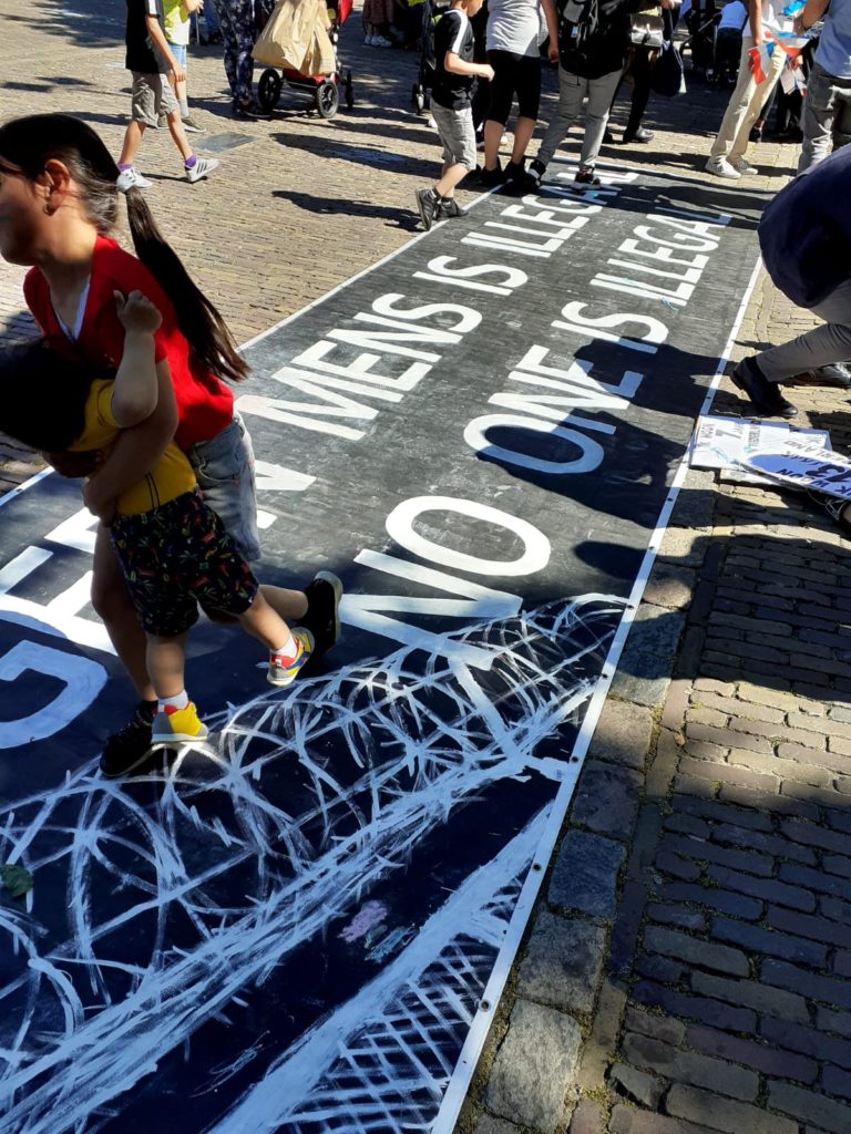 Heel groot spandoek op de grond met de tekst "GEEN MENS IS ILLEGAAL / NO ONE IS ILLEGAL" met erbij een tekening van een hek met prikkeldraad erboven.