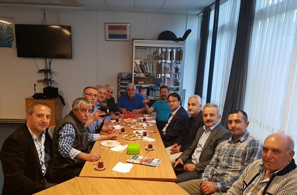 Een groep mensen aan een tafel kijken richting de camera. Onder hen zijn Halil Koçak en de Nederlandse opper-Grijze Wolf Murat Gedik. Helemaal linksboven in deze ruimte in het Beverwijkse clubhuis een afbeelding van MHP-leider Türkeş. 
