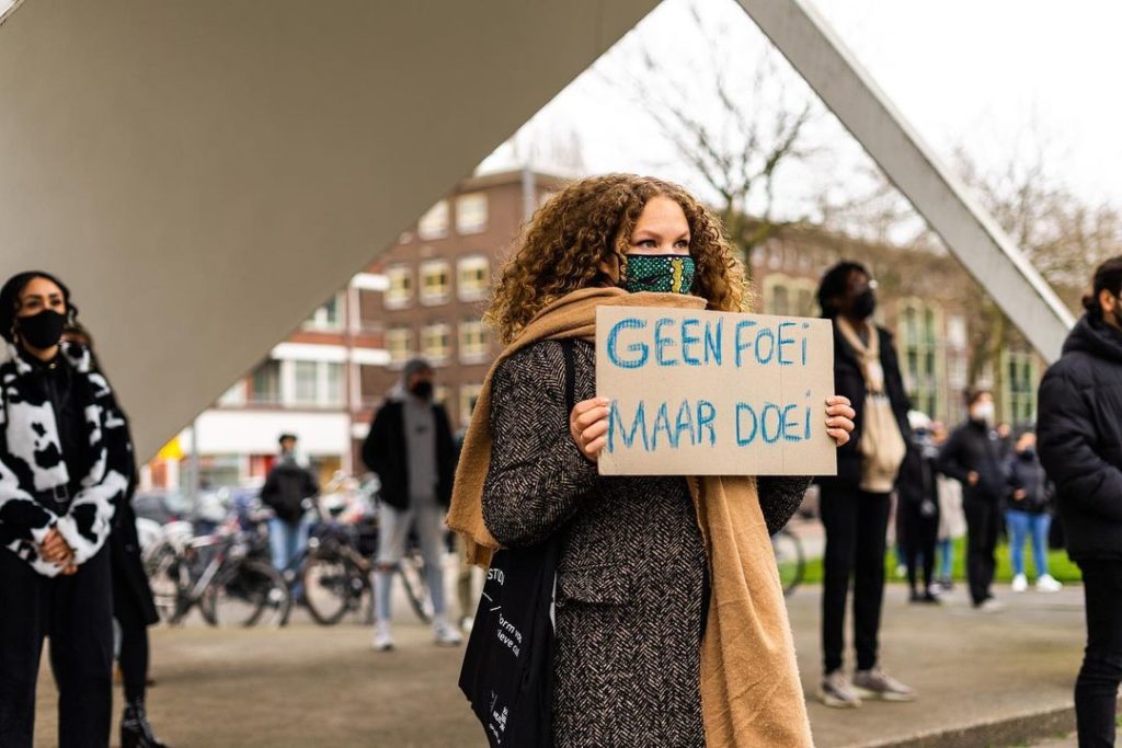 Een persoon met een grote bos lichte krullen, een groen mondkapje en een zwartwitte wollen jas aan draagt een bordje met de tekst 'Geen foei maar doei'.