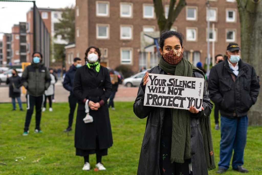 Een persoon met zwarte haren, een donkere jas en een donkerrood mondkapje draagt een bordje met de tekst "Your silence will not protect you".