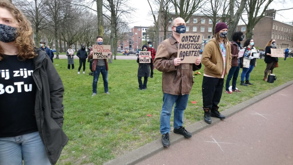 Een persoon met een bruine leren jas en een spijkerbroek draagt een bordje met de tekst "Ontsla racistische agenten". Om de persoon heen staan op een afstandje nog andere mensen met bordjes, waaronder "Geen woorden maar daden!" en "Racisme ontkennen = racisme".