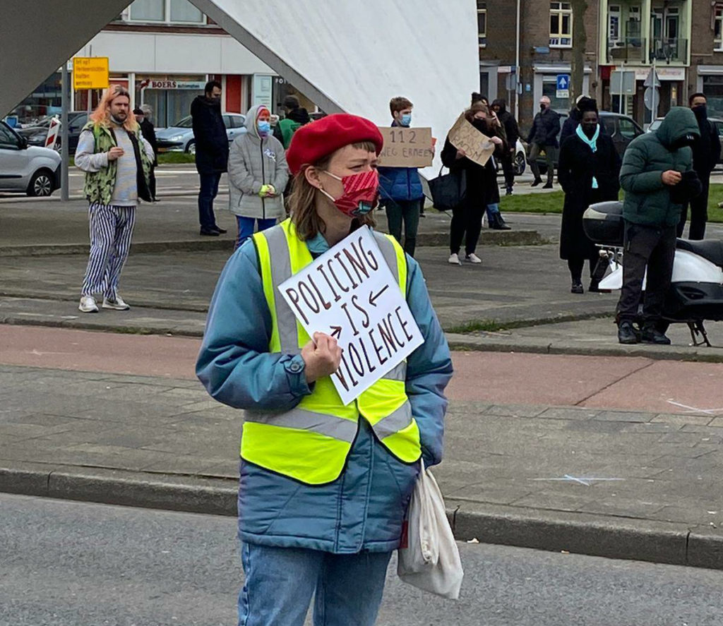 Een demonstrant met een rode baret en een veiligheidshesje draagt een bordje met de tekst "Policing is violence".