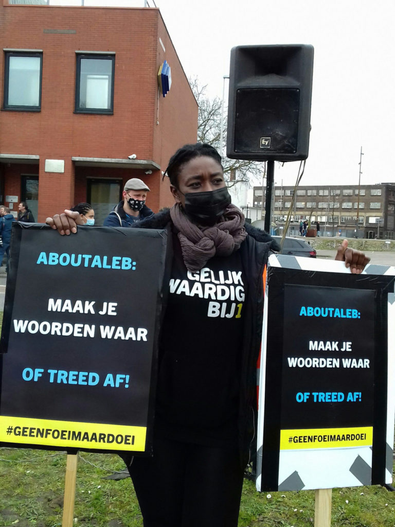 Een demonstrant met een trui van de politieke partij BIJ1 draagt twee borden met beide dezelfde tekst: "Aboutaleb: maak je woorden waar of treed af!"